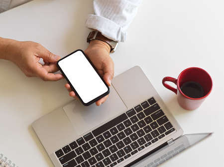 Top view of male holding smartphone with mock up screen in his hands while sitting at workspace with...