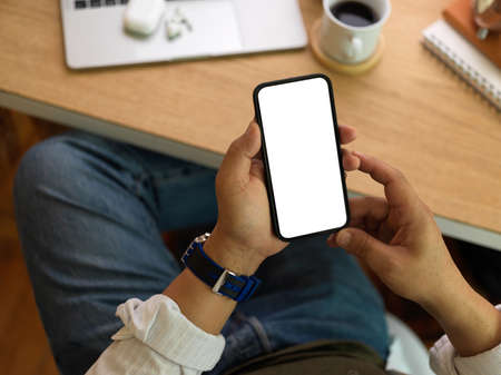 Overhead shot of businessman using smartphone with mock up screen in his hands above workspace