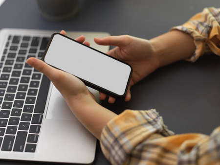 Cropped shot of female freelancer using smartphone with mock up screen in her hands while working with laptop on the tableの写真素材