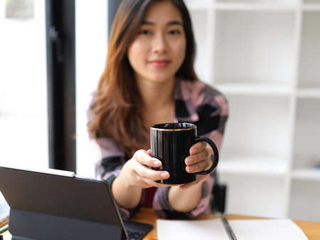 Portrait of pretty young female holding coffee cup and looking at the camera while working in the office roomの写真素材
