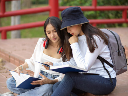 Portrait of two female students relaxed sitting on staircase and reading book together in universityの写真素材