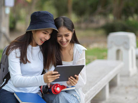 Portrait of happy two young pretty female university students using digital tablet and talking to each other while relaxing at the park in universityの写真素材