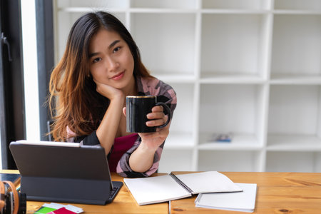 Portrait of female teenager smiling to camera and holding coffee cup while relaxing at study tableの写真素材