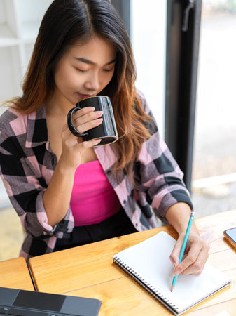 Portrait of young female freelancer drinking coffee and writing on blank notebook while working in cafeの写真素材
