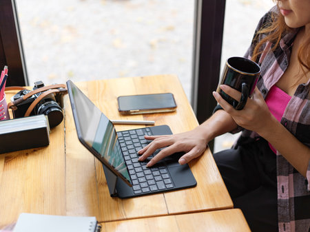 Side view of young female freelancer holding coffee cup and working with digital tablet on wooden table in cafeの写真素材