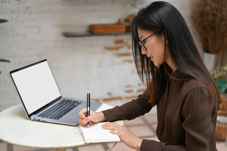 Side view of female freelancer hand writing on notebook while working with mock up laptop on round table in cafe, clipping pathの写真素材
