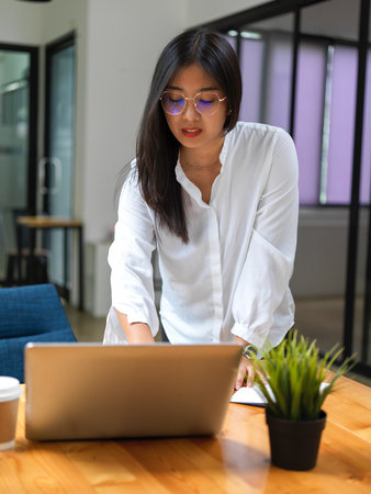 Portrait of female worker standing and working with laptop on wooden table in office roomの写真素材