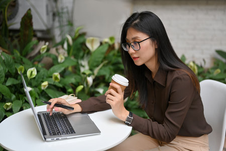 Portrait of female freelancer holding paper cup and hand typing on laptop while outdoor working in gardenの写真素材