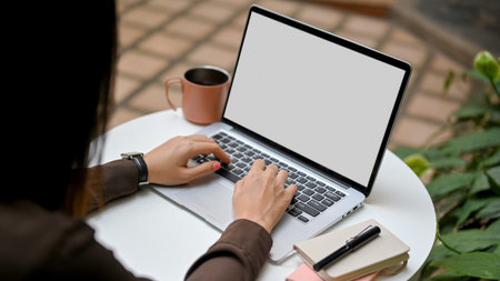 Over shoulder closeup view of female hands typing on laptop keyboard on round table in the garden at cafe, clipping pathの写真素材