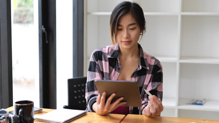 Half-length portrait of female in Scott jacket using digital tablet while sitting in libraryの写真素材