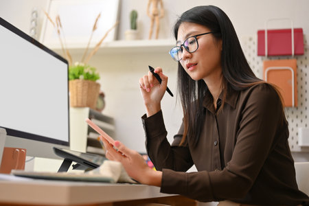 Side view of female office worker concentrating on her smartphone while working with computer in office roomの写真素材