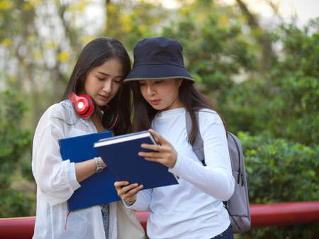 Half-length portraits of two young pretty female teenagers reading book together while standing at...