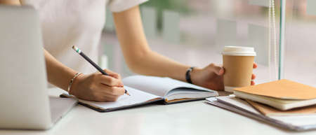 Close up view of female hand with pencil wring on notebook and holding paper cup on simple workspace