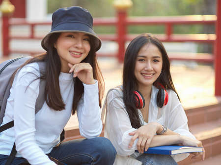 Portraits of two friendly female college students smiling to camera while relaxed sitting on staircase in the universityの写真素材