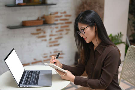 Side view of young female working with schedule book and laptop on round table in cafeの写真素材