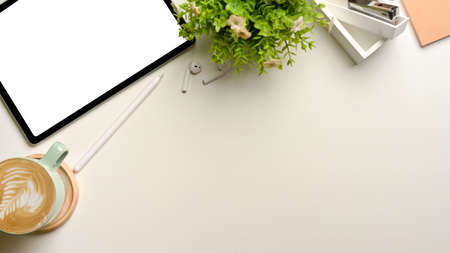 Top view of simple workspace with coffee cup, mock-up tablet, supplies, plant pot and copy space on...
