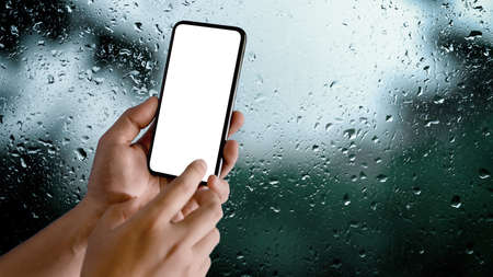 Close up view of male hands using mock-up smartphone in blurred raindrop on glass window background, clipping pathの写真素材