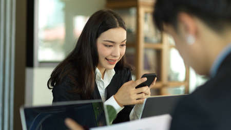 Close up view of businesswoman smiling and using smartphone while working with coworker in office roomの写真素材