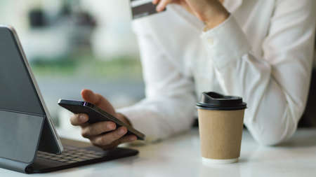 Cropped shot of female hand holding smartphone while using digital tablet on the table with paper cupの写真素材