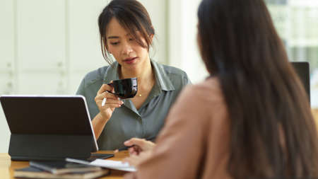 Female holding coffee cup and talking with her coworker while meeting on their projectの写真素材