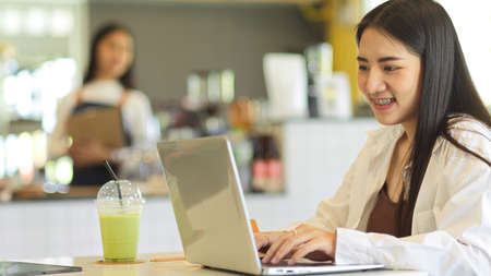 Side view of female teenager smiling and using laptop while sitting in coffee shopの写真素材