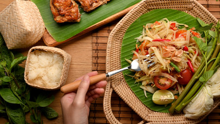 Hand with fork eating Somtum or papaya salad with sticky rice and  Thai style grilled chickenの写真素材