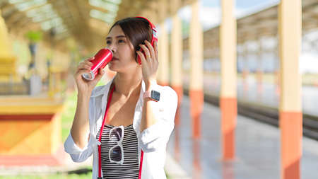 Portrait of female traveller with headphones drinking beverage can while walking in train stationの写真素材