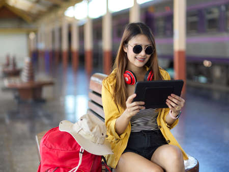 Portrait of female backpacker with sunglasses and headphone using tablet while waiting for the train at platformの写真素材