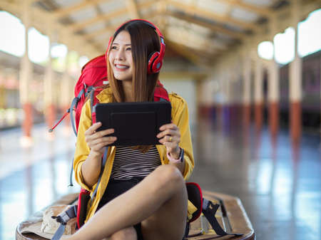 Portrait of female backpacker with headphone, bag and using digital tablet waiting the train at platformの写真素材