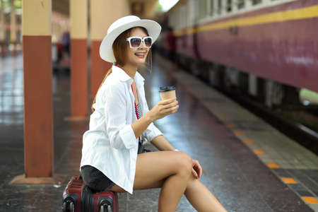 Side view of female traveler with hat and sunglasses holding paper cup while sitting at train stationの写真素材