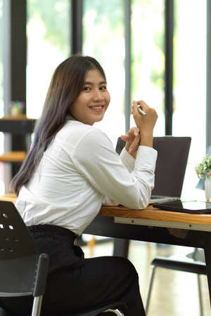 Female worker looking back and smiling to camera while sitting at workspace in cafeの写真素材