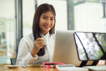 Young female office worker smiling to camera while holding coffee cup and working with laptop in cafeの写真素材