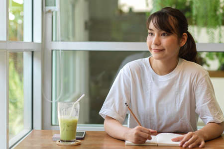Portrait of friendly female teenager looking out of the window while doing home work in cafeの写真素材
