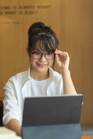 Portrait of female student with eyeglasses doing assignment on digital tablet in cafeの写真素材