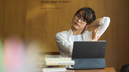 Young female student relaxing and stretching her neck while doing assignment in libraryの写真素材