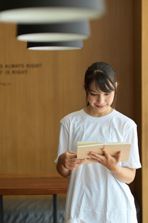 Quarter-length portrait of young female student reading a book while standing in libraryの写真素材