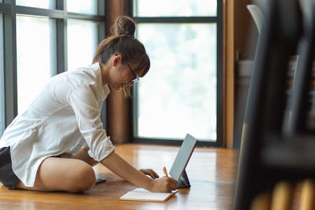 Side view of female student doing home work with notebook and digital tablet on the floor in living roomの写真素材