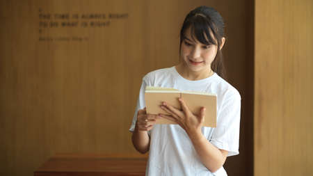 Half-length portrait of young female student reading a book while standing in libraryの写真素材