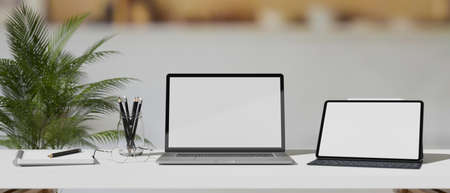 Laptop and digital tablet with mock-up screen on white table with stationery in blurred background,...