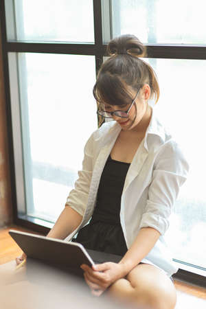 Portrait of young female student with eyeglasses looking on digital tablet while sitting on the floorの写真素材
