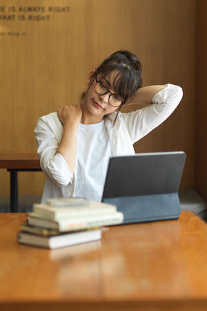 Portrait of female student relaxing and stretching her neck while preparing for exam in libraryの写真素材