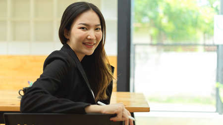Portrait of businesswoman in black suit looking back and smiling to camera while sitting in workplaceの写真素材
