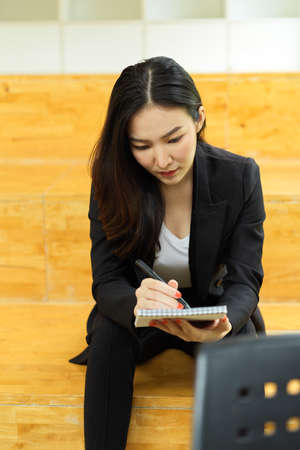 Portrait of businesswoman in black suit handwriting on schedule book while siting at stair case in office roomの写真素材