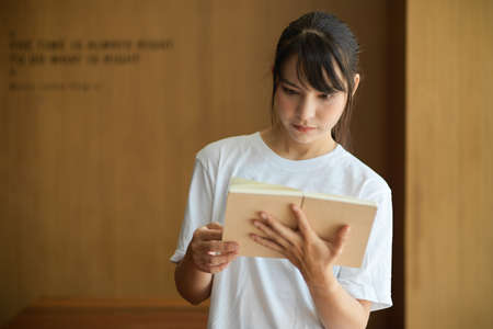 Half-length portrait of young female student reading a book while standing in libraryの写真素材