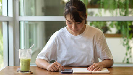 Portrait of young female teenager using smartphone and writing an idea on a notebook in cafeの写真素材