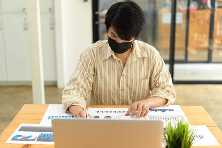 Portrait of businessman with face mask working with paperwork and laptop in office roomの写真素材