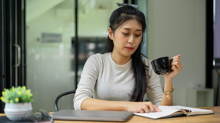 Portrait of young female student holding coffee cup while reading book to prepare for the examの写真素材