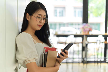 Half-length portrait young female student using smartphone and holding books while standing in cafeの写真素材