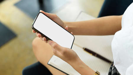 Cropped shot of female holding smartphone with mock-up screen in her hands while sitting in workplaceの写真素材