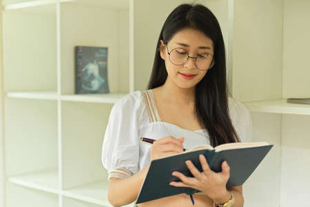 Half-length portrait of young female with eyeglasses reading book while standing in workspaceの写真素材